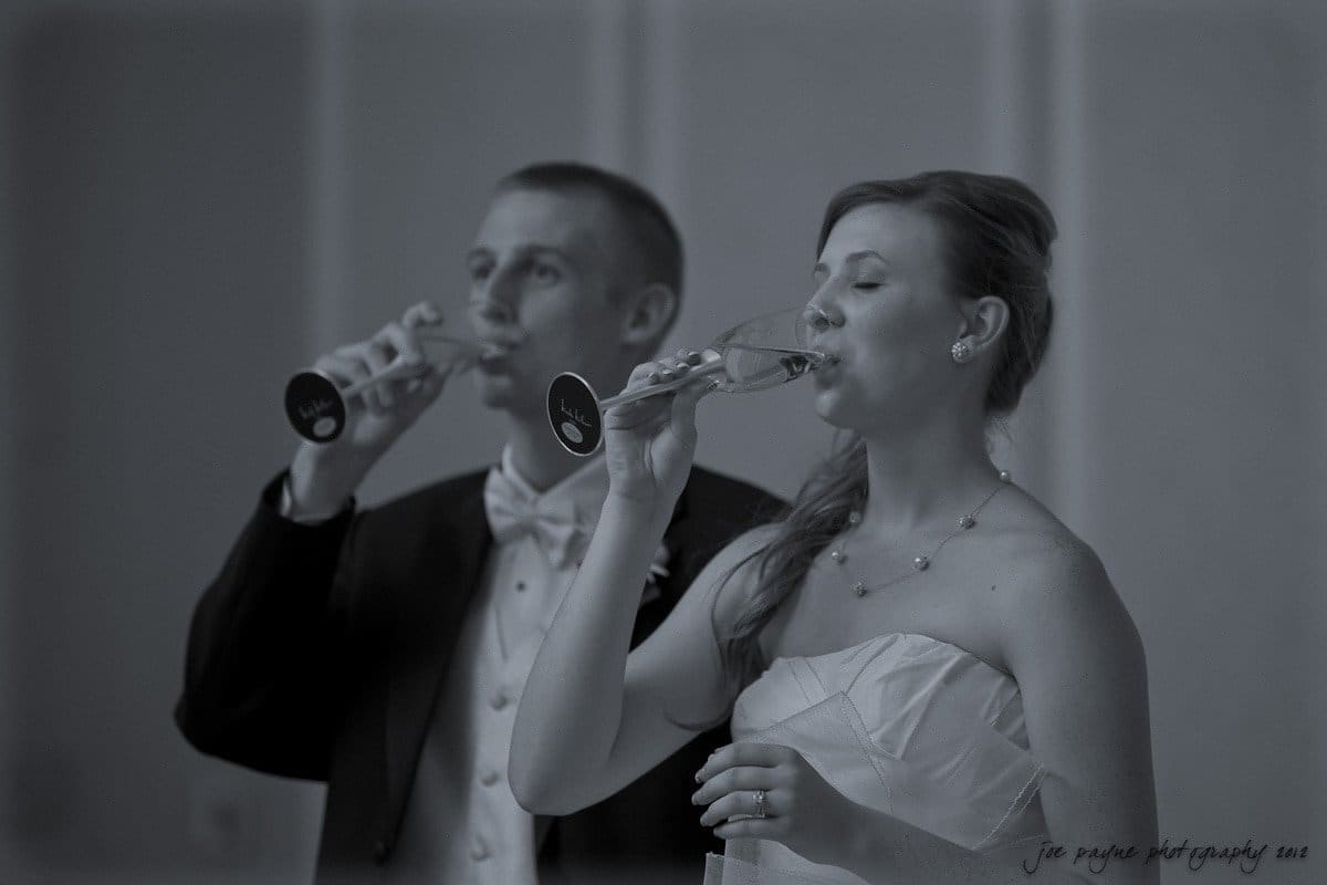 carolina inn wedding photography ~ danielle and thomas A bride and groom in formal attire sip champagne from clear glasses. The groom wears a tuxedo with a bow tie, and the bride wears a strapless dress with a necklace. Both have thoughtful expressions in a softly lit setting.