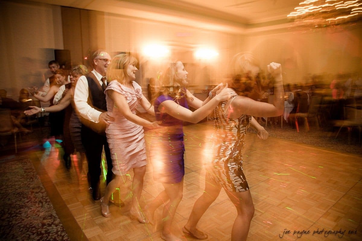 carolina inn wedding photography ~ danielle and thomas A group of people dancing in a conga line at a dimly lit party, with blurred lights creating a dynamic effect. The room is filled with tables and chairs, and a chandelier is visible above.