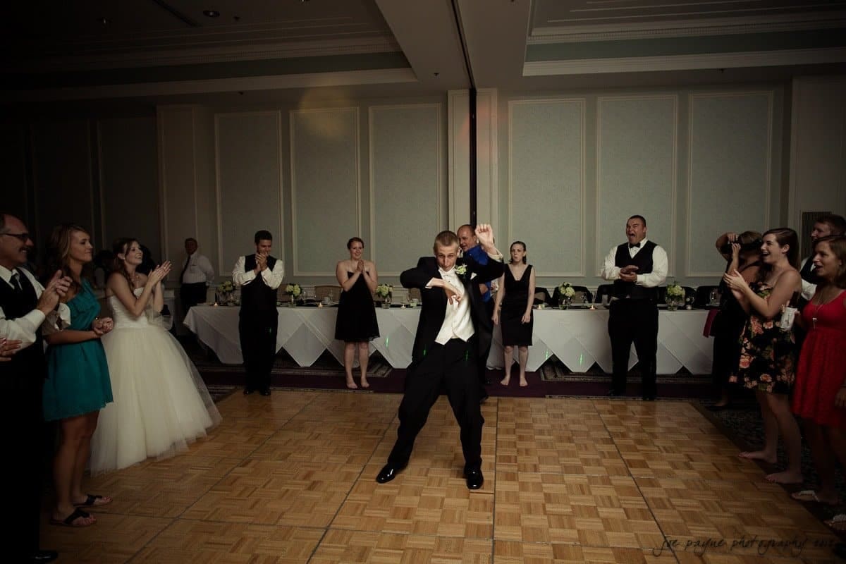 carolina inn wedding photography ~ danielle and thomas A man lifts another man on the dance floor, surrounded by applauding guests, during a lively celebration in an elegantly decorated venue. Tables with white linens and floral arrangements are visible in the background.