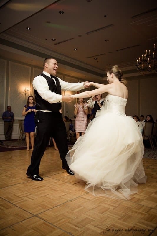 carolina inn wedding photography ~ danielle and thomas A bride in a white gown and a groom in a black vest and pants dance joyfully on a parquet floor at a wedding reception. Guests watch in the background, some clapping. The room has elegant chandeliers and light-colored walls.