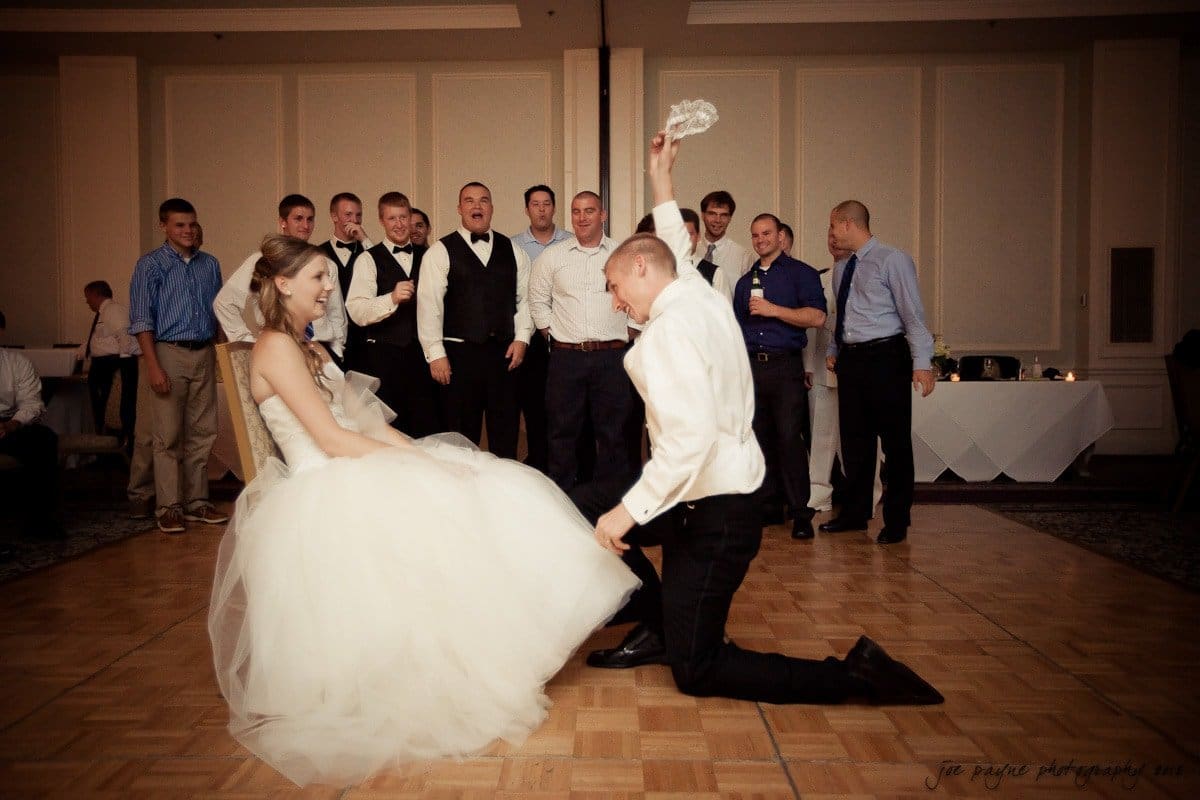 carolina inn wedding photography ~ danielle and thomas A groom playfully removes a garter from the brides leg during a wedding reception. The bride smiles while seated, and guests in formal attire watch and cheer in the background. The event takes place in a spacious, elegant hall.