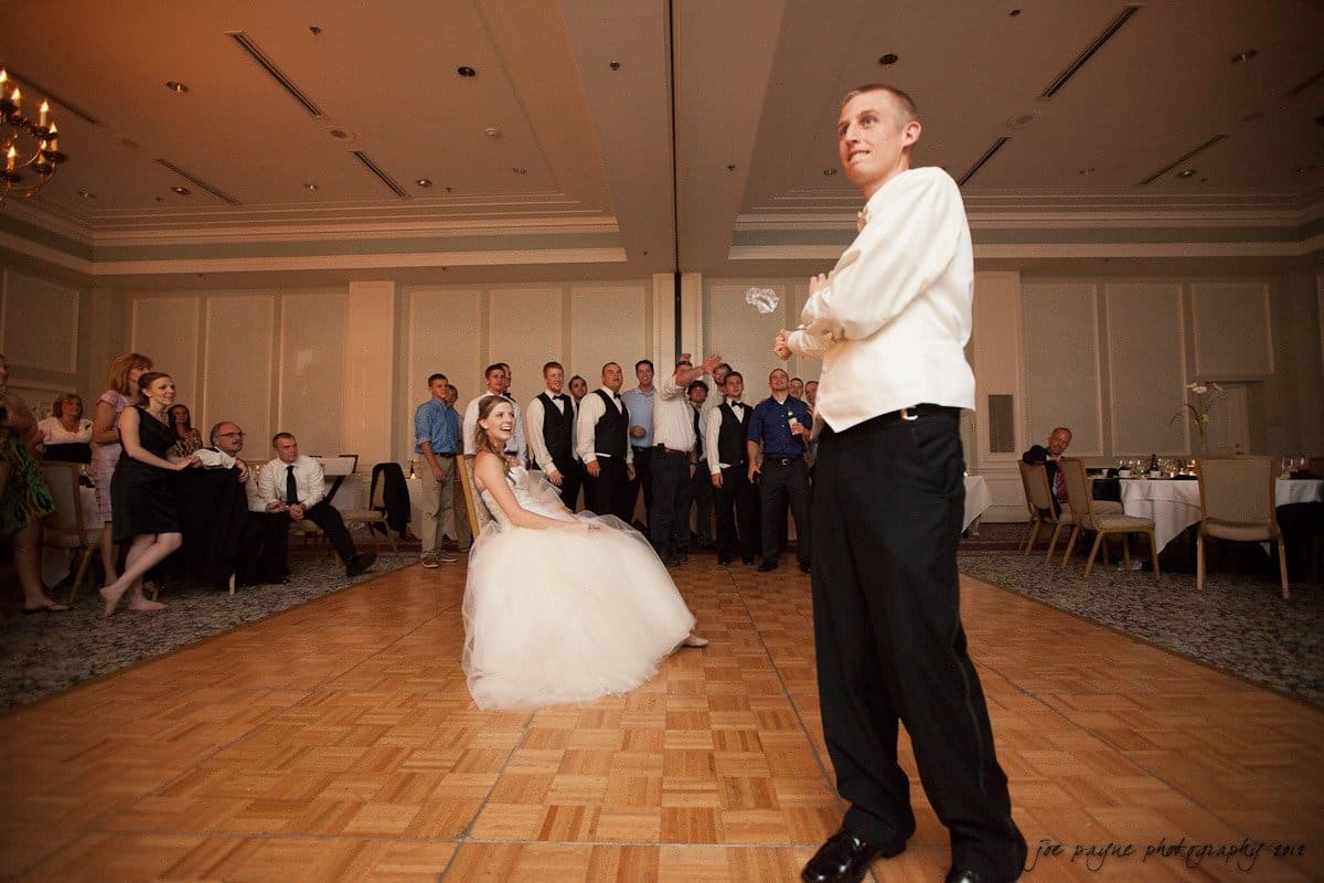 carolina inn wedding photography ~ danielle and thomas A bride sitting on a chair in a wedding dress looks expectantly at the groom, who stands with his back to a group of guests. The groom holds a garter, poised to toss it to the group in a reception hall.