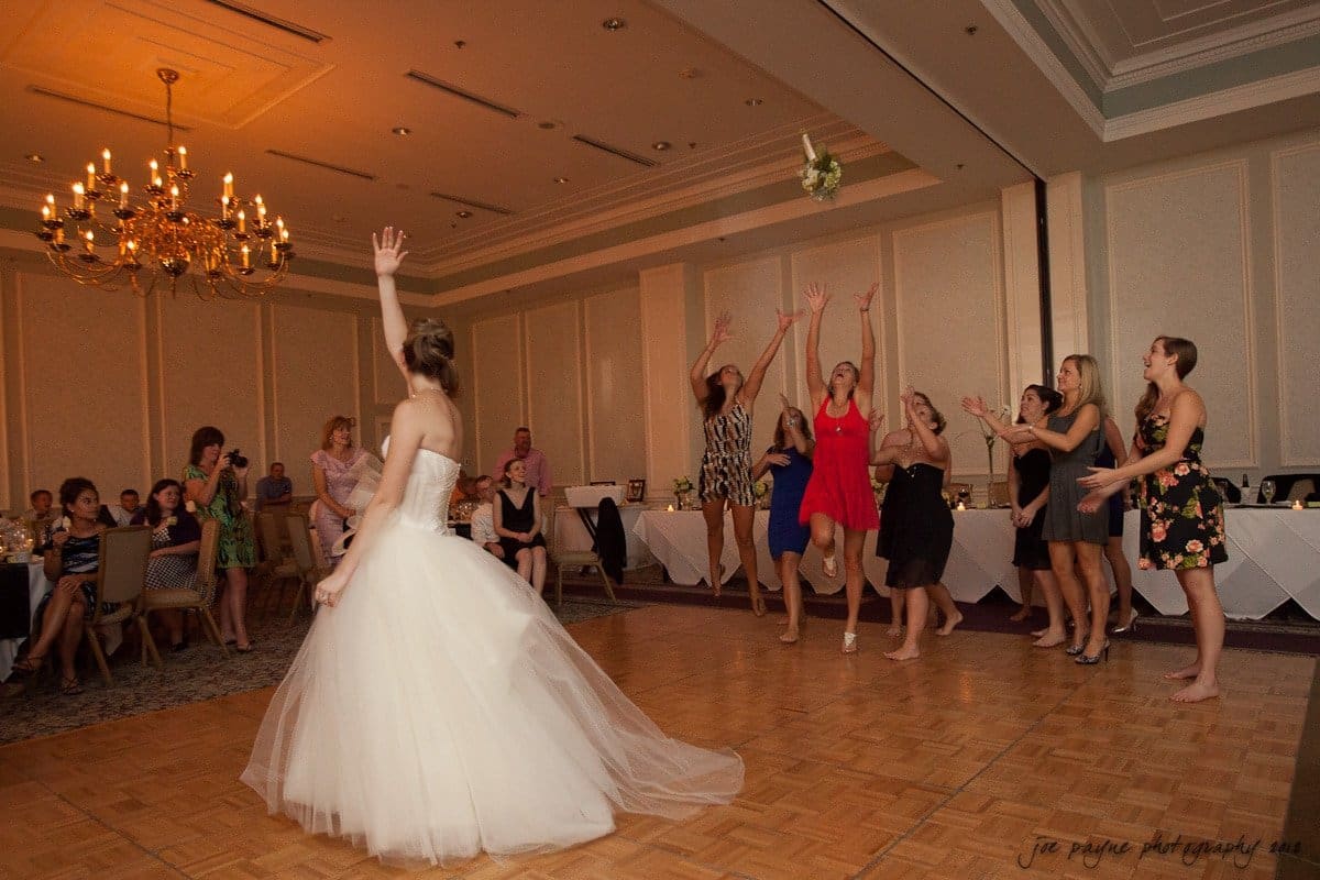 carolina inn wedding photography ~ danielle and thomas A bride in a white dress stands on a dance floor, tossing her bouquet over her shoulder. A group of women with raised arms eagerly attempt to catch it. The setting is a banquet hall with a chandelier above.