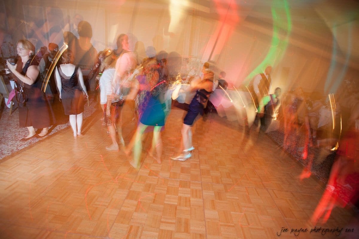 carolina inn wedding photography ~ danielle and thomas A lively dance scene on a wooden floor, with people blurred in motion, creating colorful light trails. The background shows a warmly lit room filled with activity and energy.