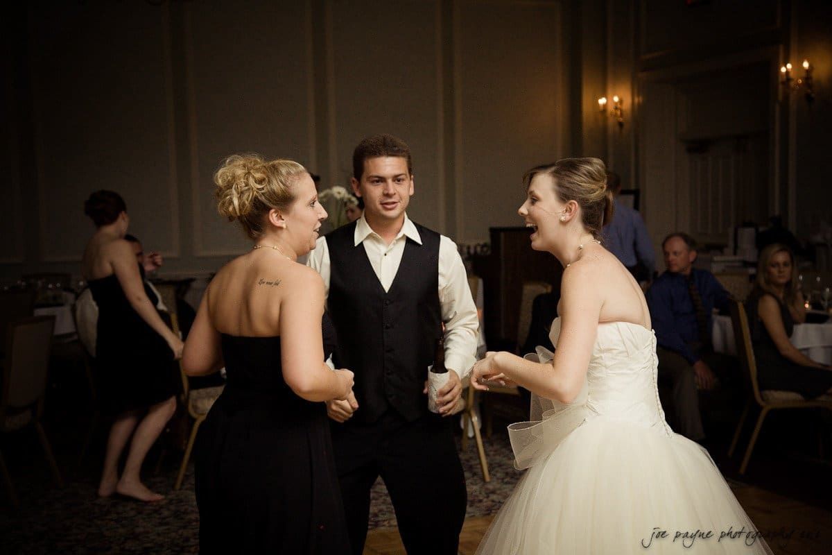 carolina inn wedding photography ~ danielle and thomas Three people are joyfully dancing together at a wedding reception. Two women, one in a black dress and the other in a white wedding gown, are smiling, while a man in a vest and tie stands between them. The room is dimly lit, with elegant decor and guests seated in the background.