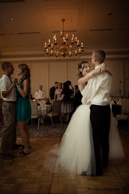 carolina inn wedding photography ~ danielle and thomas Bride and groom dance together in a warmly lit ballroom, while another couple dances nearby. A chandelier hangs overhead, and guests mingle in the background.