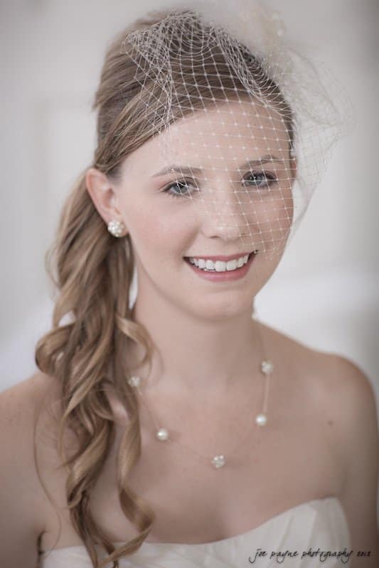 carolina inn wedding photography ~ danielle and thomas A smiling bride wearing a white dress and pearl jewelry. Her hair is styled with loose waves and covered with a birdcage veil. The background is softly blurred.