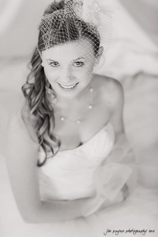 carolina inn wedding photography ~ danielle and thomas A bride with curled hair and a small veil smiles while wearing a white strapless gown. The black and white image captures her looking up, showing delicate jewelry and soft fabric details.