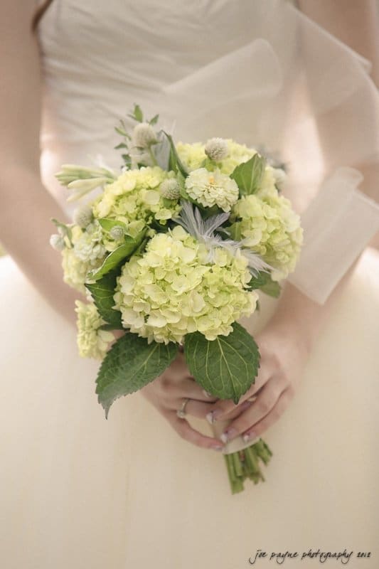 carolina inn wedding photography ~ danielle and thomas A bride holds a bouquet of pale green and white flowers, featuring hydrangeas, roses, and greenery, against her white dress. The image has a soft and dreamy quality.