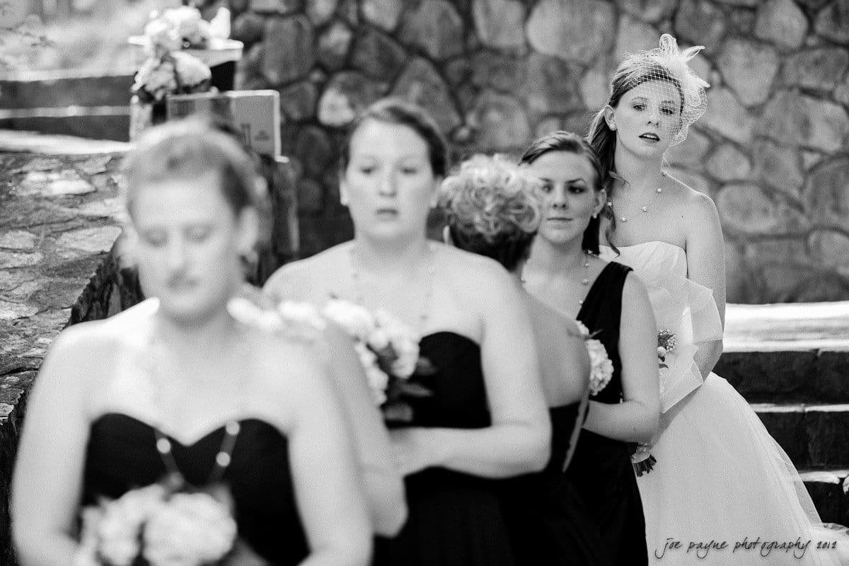 carolina inn wedding photography ~ danielle and thomas A black and white photo of a bride and bridesmaids. The bride stands slightly apart in a strapless gown with a veil. The bridesmaids, in darker dresses, hold bouquets as they proceed in a line. Stone steps and a wall form the background.