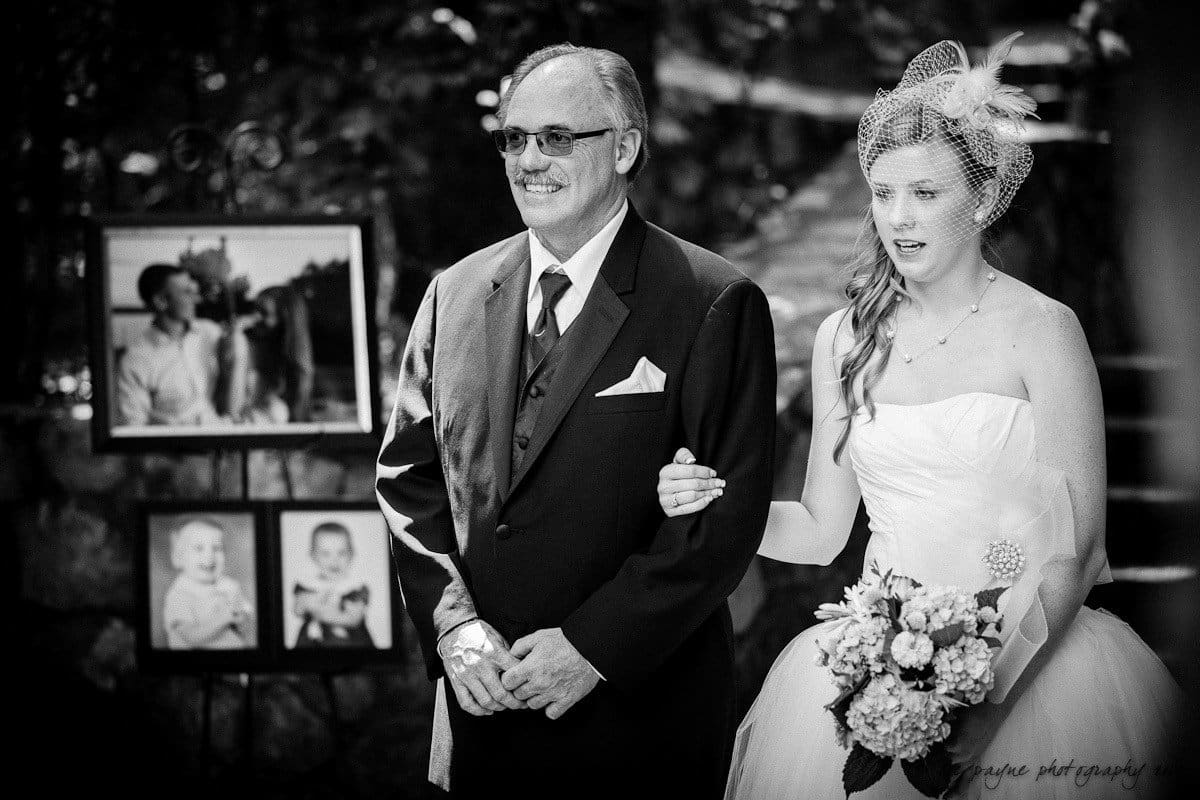 carolina inn wedding photography ~ danielle and thomas A bride in a white gown and veil walks arm-in-arm with a man in a suit, likely her father. They are outdoors, with framed photos of children displayed in the background. The scene conveys a wedding atmosphere.