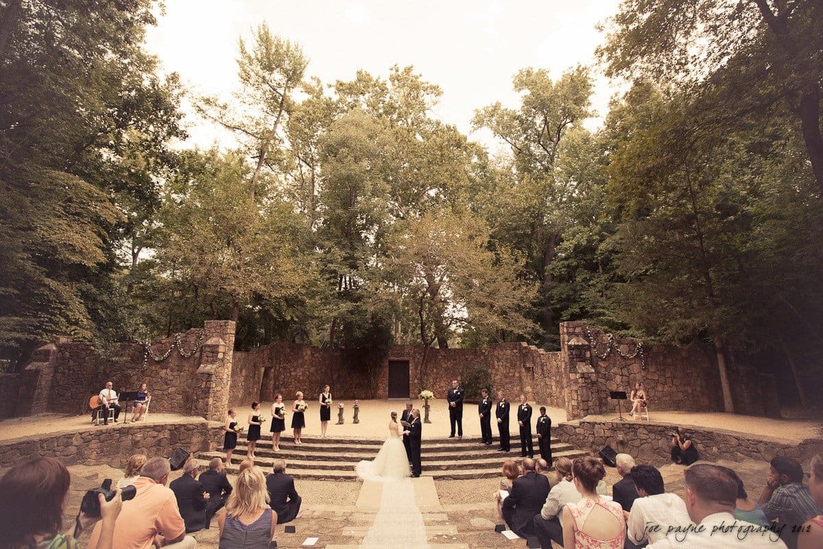 carolina inn wedding photography ~ danielle and thomas An outdoor wedding ceremony takes place in a forested stone amphitheater. The bride and groom stand at the center surrounded by their bridal party, with seated guests observing. Tall trees and rustic stone walls add to the natural setting.