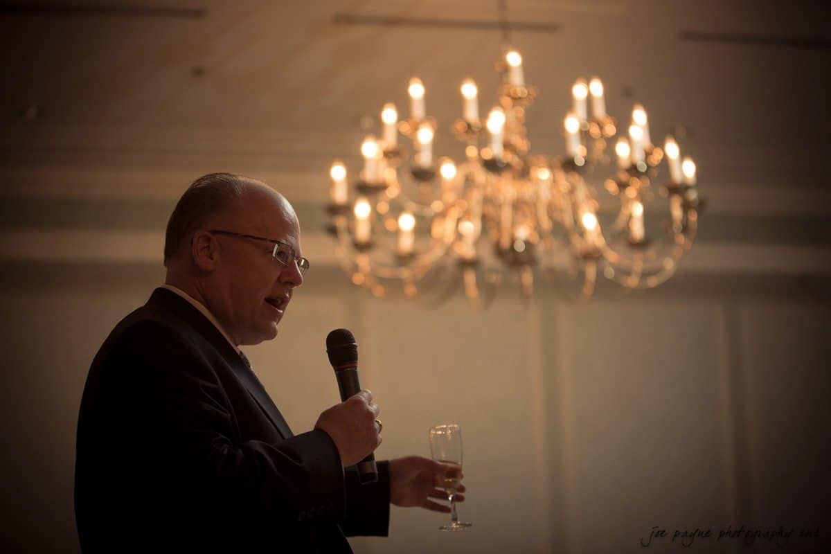 A man in a suit speaks into a microphone while holding a glass, standing under a lit chandelier in a softly lit room.