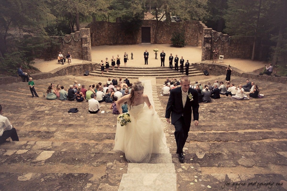 carolina inn wedding photography ~ danielle and thomas A bride and groom ascend stone steps at an outdoor amphitheater, holding hands. The wedding party and guests are seated in a semicircle, surrounded by trees and a rustic stone backdrop. The scene exudes a serene and romantic atmosphere.