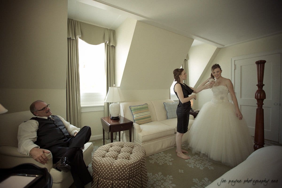 carolina inn wedding photography ~ danielle and thomas A bride in a white gown is being assisted by a woman in a black dress in a warmly lit room. A man in a suit sits on a couch, watching them. The space is decorated with soft furnishings and a window lets in natural light.