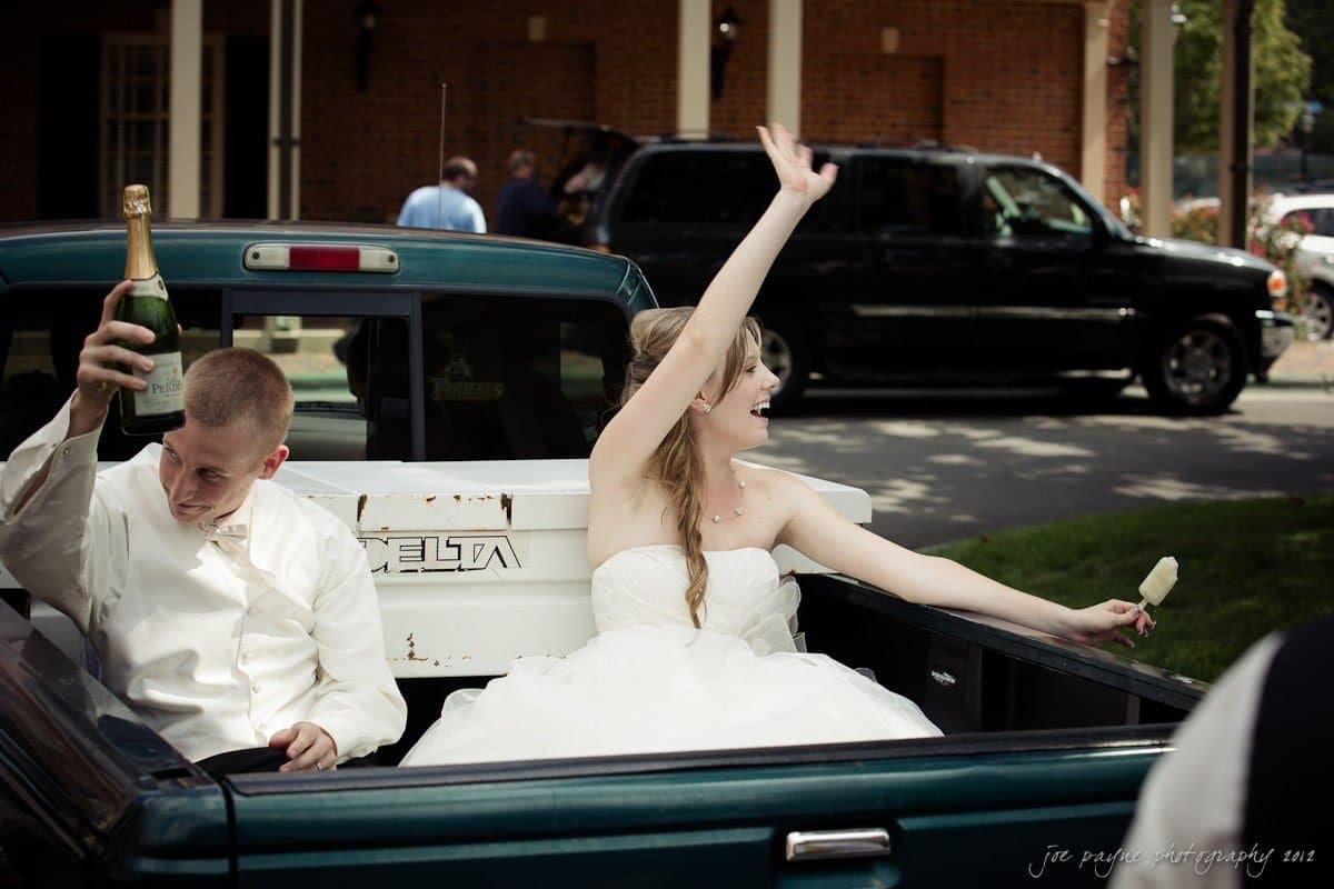carolina inn wedding photography ~ danielle and thomas A bride and groom in wedding attire sit in the back of a pickup truck. The groom holds a bottle of champagne, while the bride waves enthusiastically and holds an ice pop. The truck is parked in a driveway with people and cars in the background.