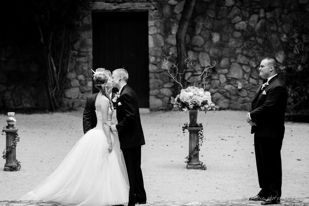 carolina inn wedding photography ~ danielle and thomas A couple stands at an outdoor wedding ceremony, sharing a kiss. The bride wears a flowing white gown, and the groom is in a dark suit. A simple floral arrangement is displayed nearby, and an officiant watches the couple. Stone walls are visible in the background.