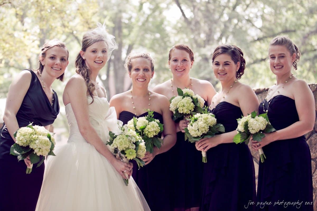 carolina inn wedding photography ~ danielle and thomas Bride in a white gown and veil stands with five bridesmaids in black dresses holding bouquets of white and green flowers. They are outdoors, smiling, with greenery in the background.