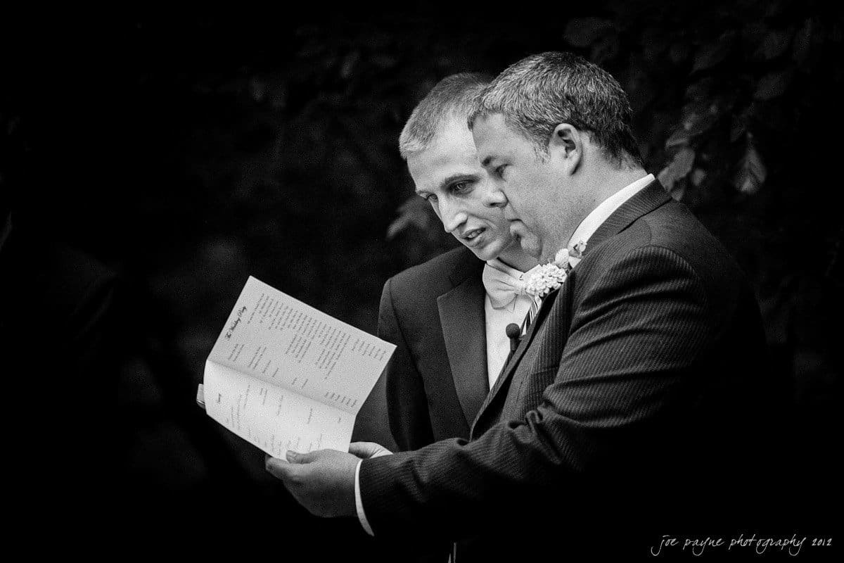 carolina inn wedding photography ~ danielle and thomas Two men dressed in formal suits are closely reading from a sheet of paper. One man is pointing at the text while the other looks on, both appearing focused. The background is dark, highlighting the two men.