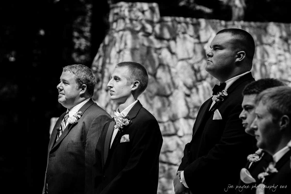 carolina inn wedding photography ~ danielle and thomas A group of men in formal suits, with flower boutonnieres, stand in a row during a ceremony. The background features a stone wall. The image is in black and white.