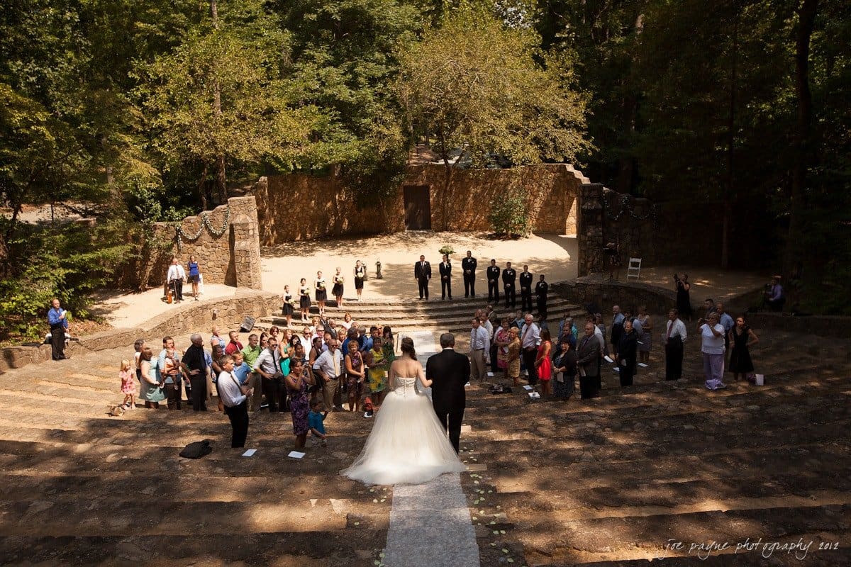 carolina inn wedding photography ~ danielle and thomas A bride and groom stand at the altar in an outdoor amphitheater surrounded by guests. The sunlit scene is framed by lush trees, with attendees seated on stone steps. The ceremony takes place under a clear sky.