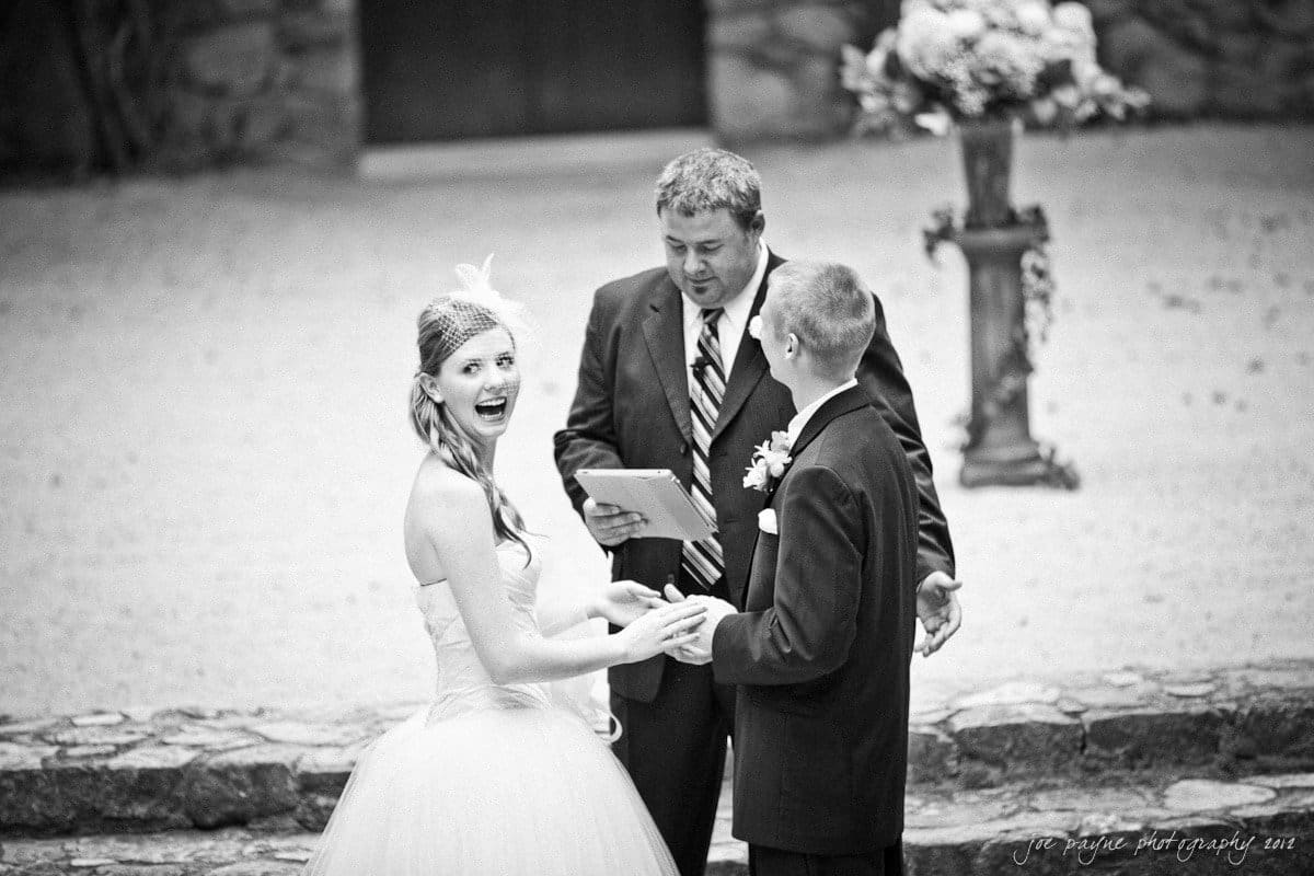 carolina inn wedding photography ~ danielle and thomas A black and white photo of a bride and groom holding hands in front of an officiant during a wedding ceremony. The bride is smiling brightly at the camera. The scene is outdoors with a stone floor and floral decorations in the background.
