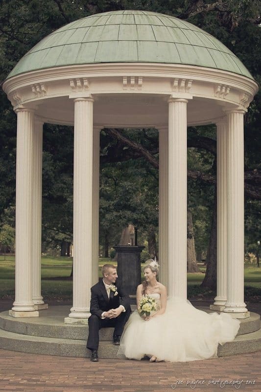 carolina inn wedding photography ~ danielle and thomas A bride in a flowing white gown sits beside a groom in a black suit under a round, white gazebo with columns. They are on a brick pathway surrounded by lush green trees. The groom holds the brides hand as they look at each other smiling.