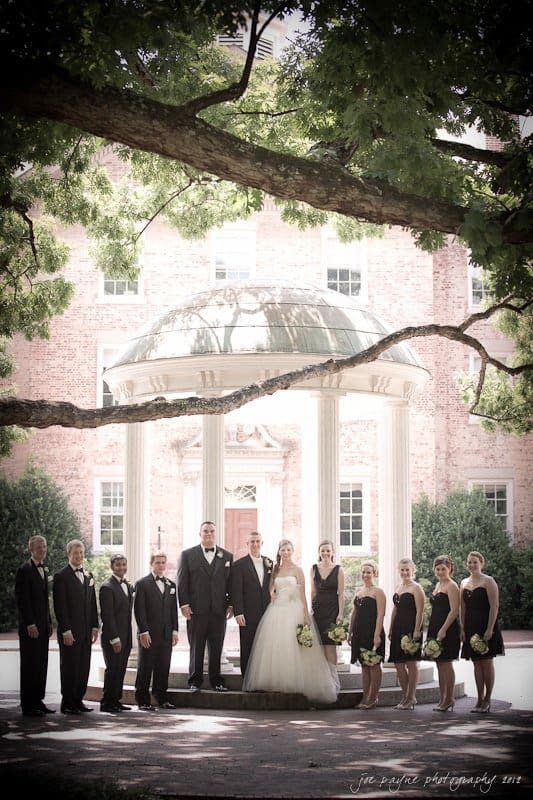 carolina inn wedding photography ~ danielle and thomas A wedding party stands under a large tree in front of a domed gazebo. The bride and groom are surrounded by bridesmaids in black dresses and groomsmen in black suits, all smiling. A brick building is visible in the background.