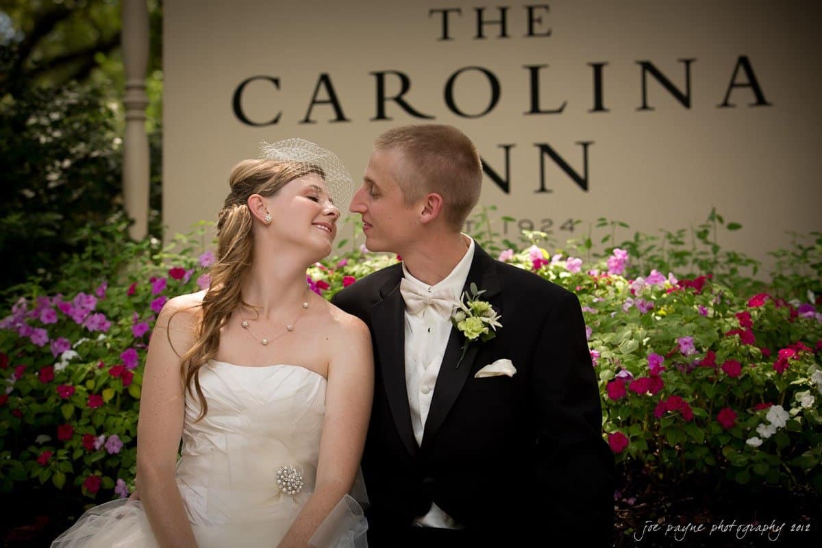 carolina inn wedding photography ~ danielle and thomas A bride and groom sit closely, touching noses and smiling in front of a sign for The Carolina Inn. The bride is in a white strapless dress, while the groom wears a black tuxedo with a white boutonniere. Theyre surrounded by colorful flowers.