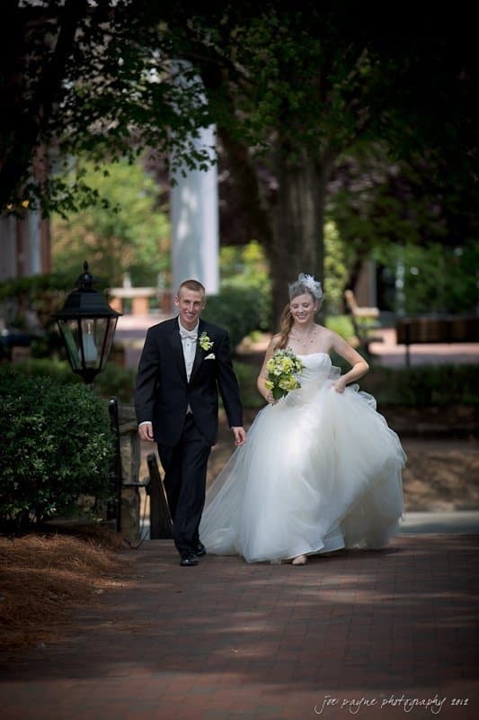 carolina inn wedding photography ~ danielle and thomas A bride and groom walk arm in arm down a brick path. The bride wears a white gown and holds a bouquet of yellow flowers. The groom wears a black suit. They smile as they walk under the shade of trees.