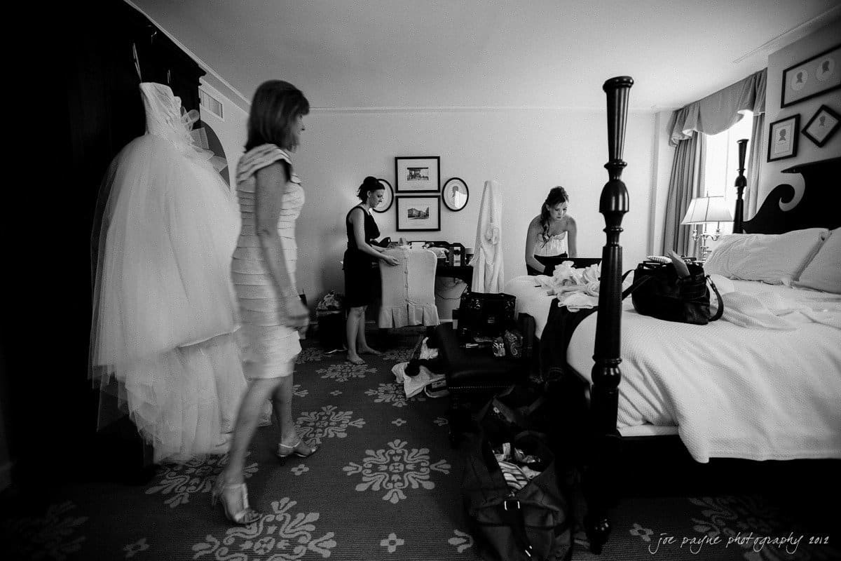 carolina inn wedding photography ~ danielle and thomas A black and white photo of a bridal preparation scene. Three women are in a cluttered room with a bed. One stands near a hanging wedding dress, another is seated on the bed, and the third is adjusting a garment.
