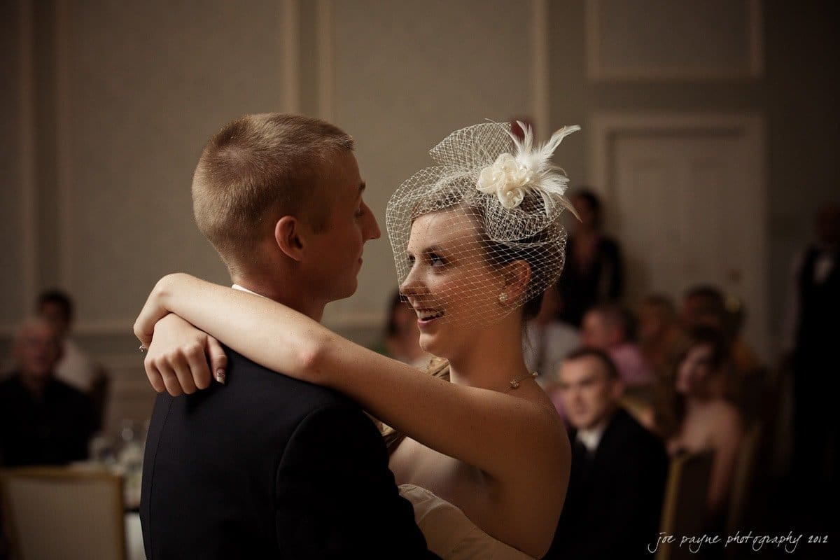 carolina inn wedding photography ~ danielle and thomas A bride and groom share their first dance at a wedding reception. The bride wears a dress with a birdcage veil and floral headpiece, while the groom is in a suit. Guests are seated in the softly lit background.
