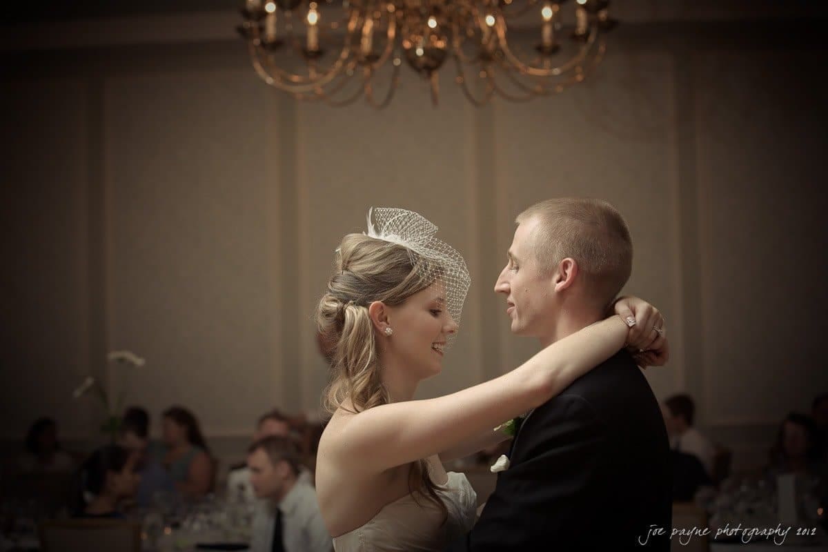 carolina inn wedding photography ~ danielle and thomas A bride and groom share a dance in a warmly lit reception hall. The bride wears a white dress with a small veil, and the groom is in a dark suit. A chandelier hangs above them, and guests are seated in the background.