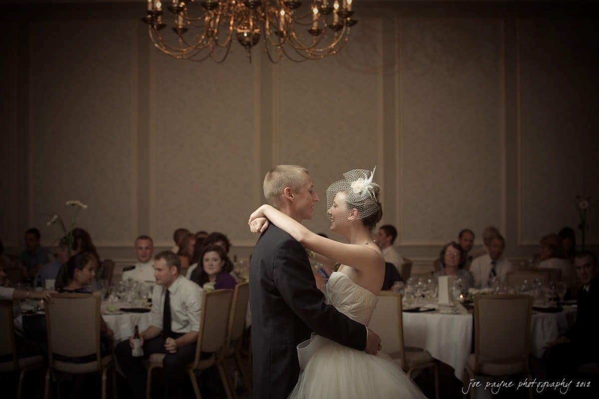 carolina inn wedding photography ~ danielle and thomas A bride and groom share their first dance in a softly lit banquet hall. Guests sit at round tables watching the couple. A chandelier hangs from the ceiling, adding elegance to the intimate setting.