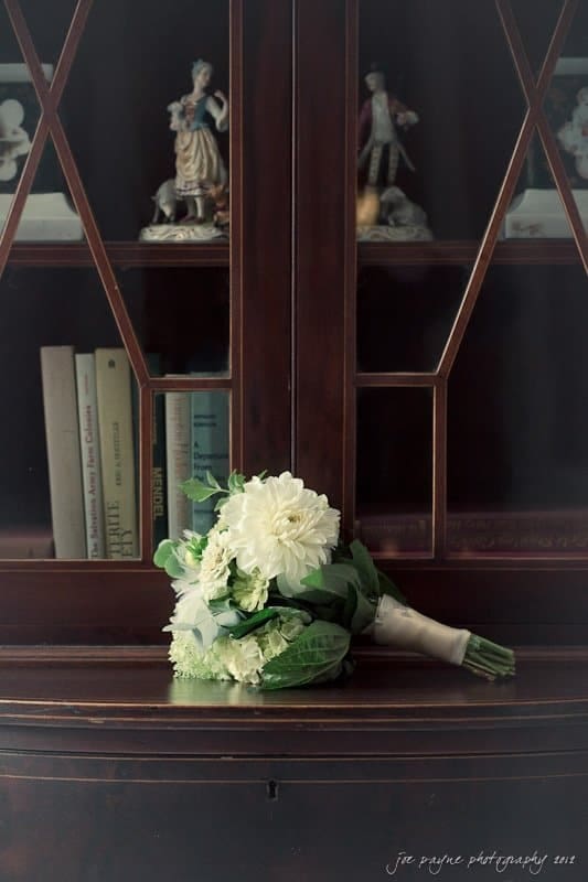 carolina inn wedding photography ~ danielle and thomas A bouquet of white flowers rests on a polished wooden cabinet with glass-paned doors. Inside the cabinet are books and decorative figurines. The scene is illuminated with soft, natural light.