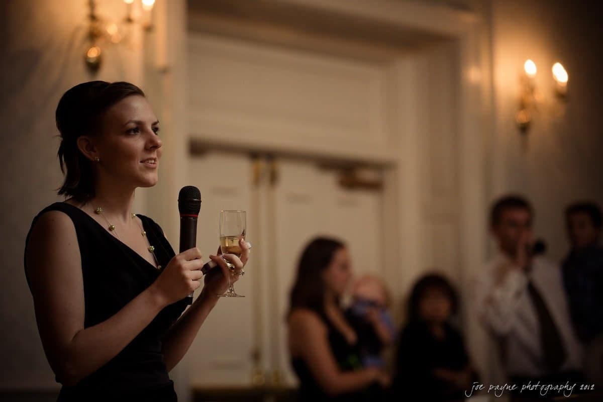 carolina inn wedding photography ~ danielle and thomas A woman in a black dress is delivering a speech while holding a microphone and a glass of champagne. She stands in a dimly lit room with blurred people in the background, suggesting an event or gathering.