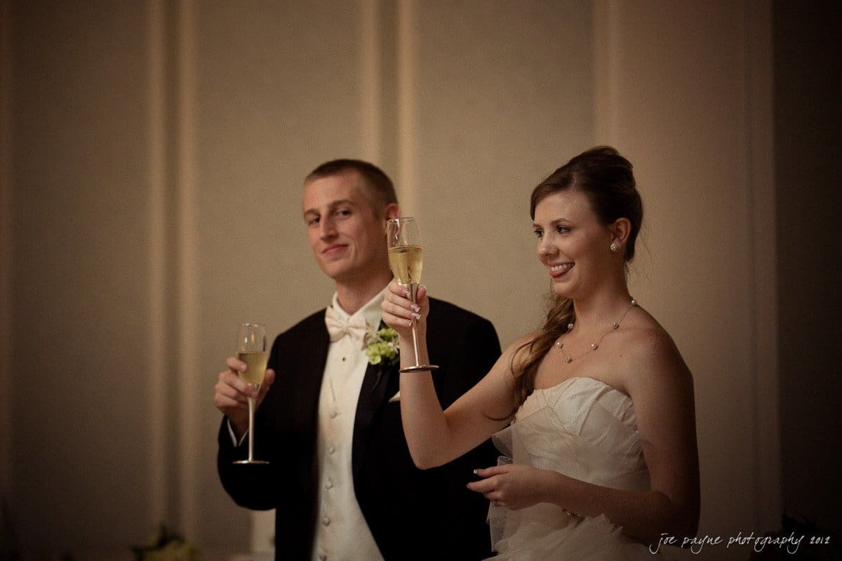 carolina inn wedding photography ~ danielle and thomas A bride and groom hold champagne glasses during a toast, smiling in a warmly lit room. The bride is wearing a strapless white gown, and the groom is in a black tuxedo with a white bow tie and boutonniere.