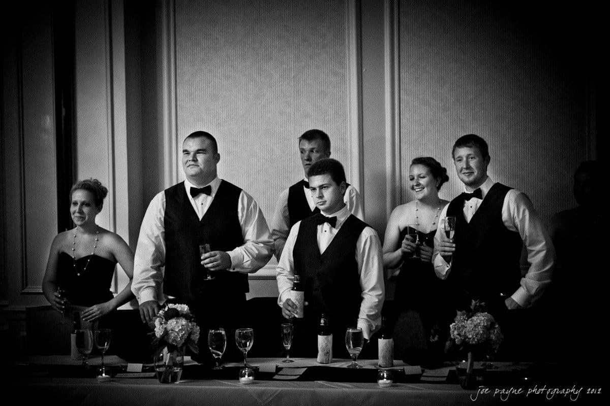 carolina inn wedding photography ~ danielle and thomas A group of wedding party members stand together, dressed in formal attire—men in vests and bow ties, women in elegant dresses. They hold glasses, likely for toasting, and smile in a warmly lit banquet setting.