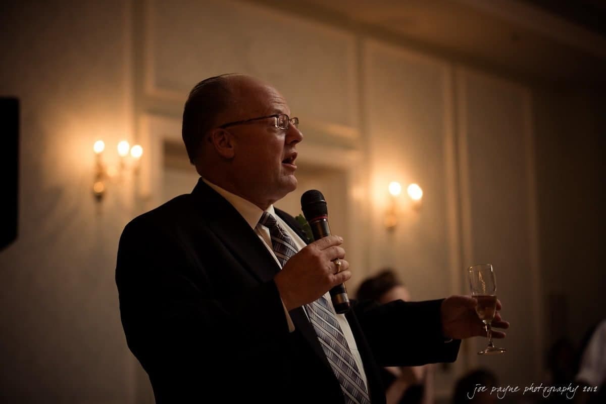 carolina inn wedding photography ~ danielle and thomas A man in a suit holds a microphone in one hand and a champagne glass in the other, speaking in a dimly lit room with ornate wall sconces.