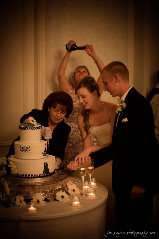 carolina inn wedding photography ~ danielle and thomas A couple cuts a tiered wedding cake decorated with black silhouettes. A woman assists them, while another person photographs the moment on a phone. The table is adorned with white flowers and candles.