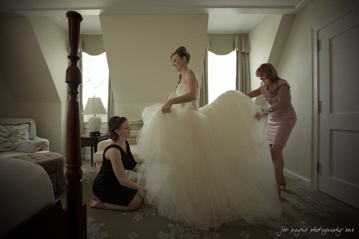 carolina inn wedding photography ~ danielle and thomas A bride stands in a room with a large, white wedding dress as two women assist her. One woman, kneeling, adjusts the dress while the other stands nearby holding a flower. The room features a bed, windows, and a cream-colored carpet.