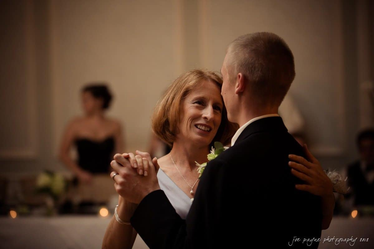 carolina inn wedding photography ~ danielle and thomas A couple dressed in formal attire share a dance in a softly lit room. The woman smiles warmly as they embrace. A blurred background with people sitting and floral decorations create an elegant ambiance.