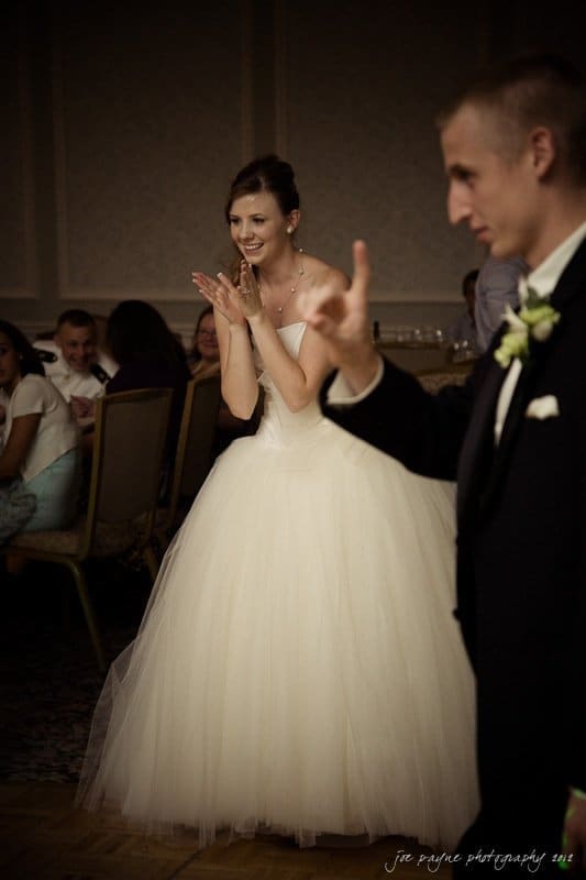 carolina inn wedding photography ~ danielle and thomas A bride in a white gown smiles and claps while standing, surrounded by guests seated at tables. A man in a suit, possibly the groom, is in the foreground, blurred. The setting is a dimly lit room, suggesting a wedding reception.