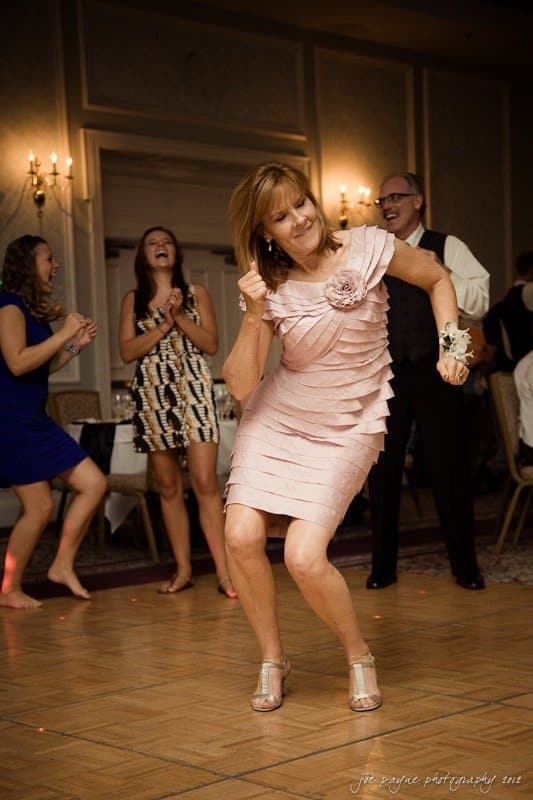 carolina inn wedding photography ~ danielle and thomas A woman in a light pink dress dances energetically on a wooden floor. She is surrounded by people who are clapping and smiling. The room is elegantly decorated with chandeliers and beige walls.