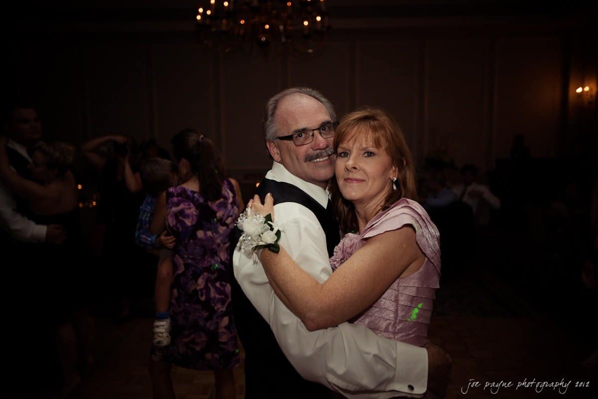 carolina inn wedding photography ~ danielle and thomas A couple dances together at a formal event. The man wears a dark suit with a vest, and the woman wears a lavender dress. Other couples are visible in the softly lit background, creating an intimate atmosphere.