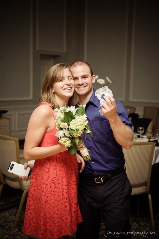 carolina inn wedding photography ~ danielle and thomas A smiling woman in a red dress holds a bouquet, and a man in a blue shirt proudly shows a garter. Theyre posing happily indoors at a celebratory event with tables and chairs in the background.