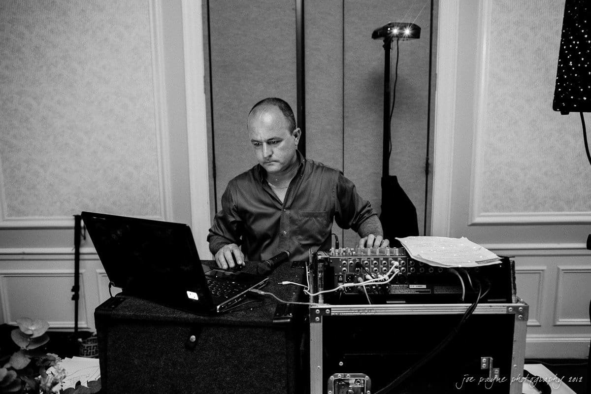 carolina inn wedding photography ~ danielle and thomas A man is focused on operating audio equipment at an event. He is using a laptop and multiple control panels, surrounded by equipment. The setting is a formally decorated room.