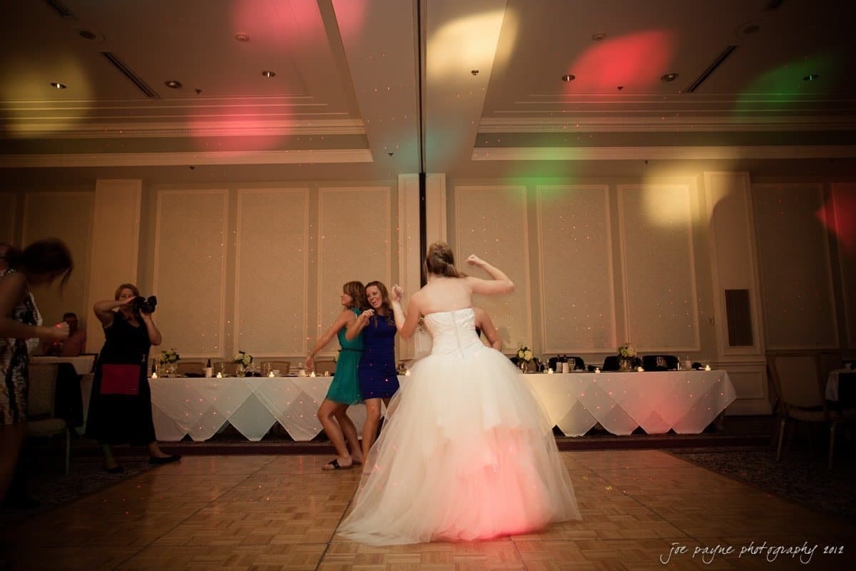 carolina inn wedding photography ~ danielle and thomas A bride in a white gown dances with guests on a dimly lit dance floor, surrounded by multicolored lights. A long table with decorations is in the background.