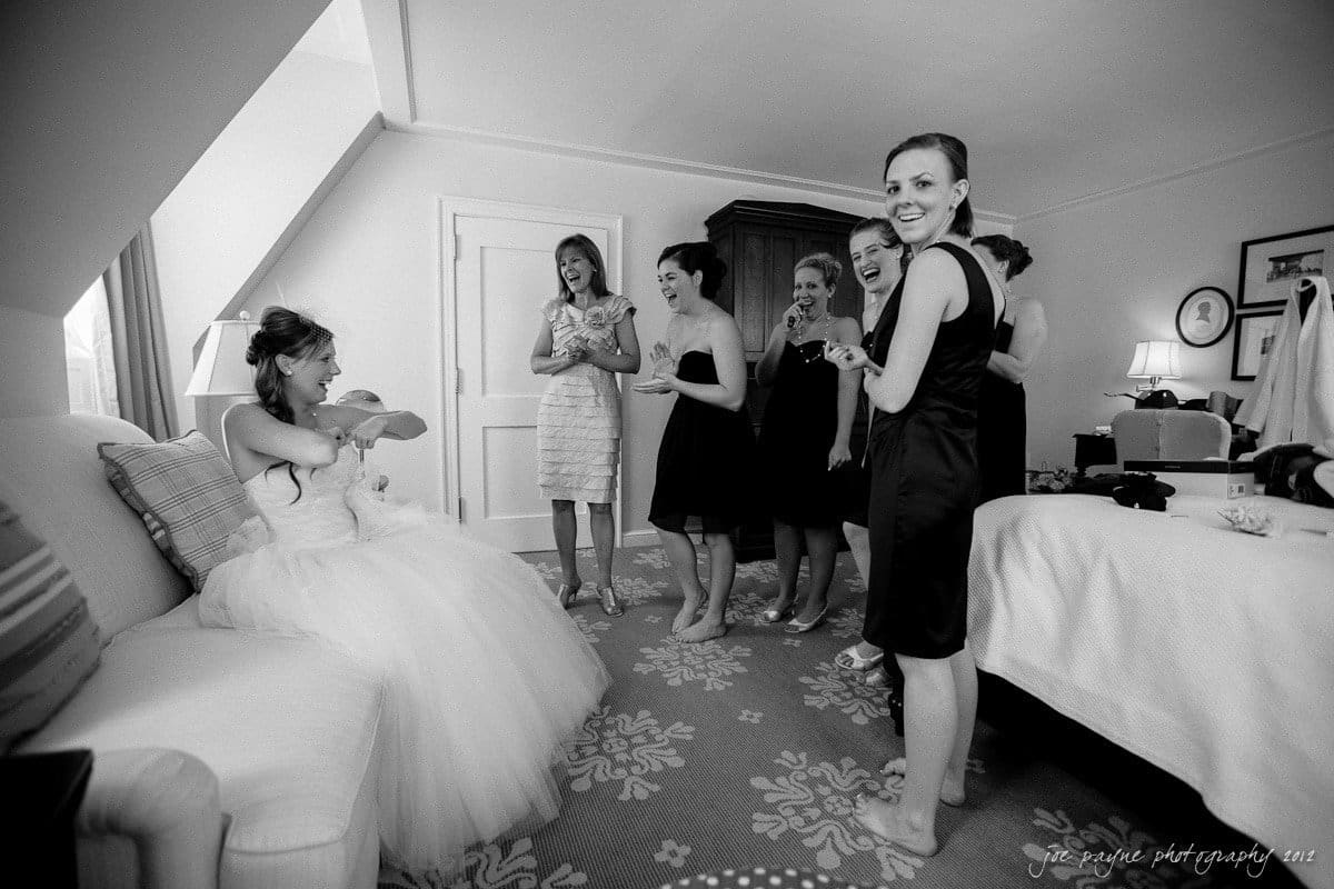 carolina inn wedding photography ~ danielle and thomas A bride in a wedding dress sits on a sofa, smiling and extending her arms toward a standing baby. Around her, a group of six women in black dresses laugh and react joyfully in a room with elegant decor.