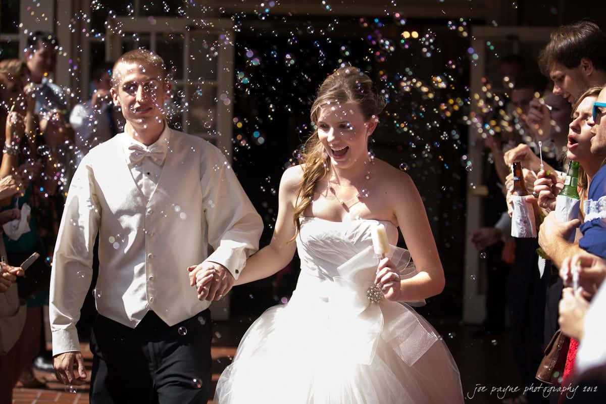 carolina inn wedding photography ~ danielle and thomas A bride and groom walk hand in hand, smiling as they are surrounded by bubbles. The groom is in a white vest and bow tie, while the bride wears a white strapless gown. Guests on either side blow bubbles, adding a festive atmosphere.
